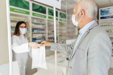 Sale of prescription drugs. A female pharmacist and dressed in a white uniform with a protective mask on his face in a plastic bag of medicines and supplements delivered to the mature male customer