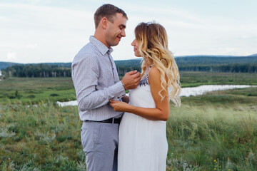 Happy young couple, expecting a baby. Man hugs his pretty pregnant woman in a white dress outdoors.