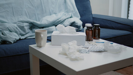 Close up of coffee table with medicine and bottle of pills. Nobody in living room with medicaments, capsules, box of tissues and thermometer. Empty space for sick patient and healing treatment