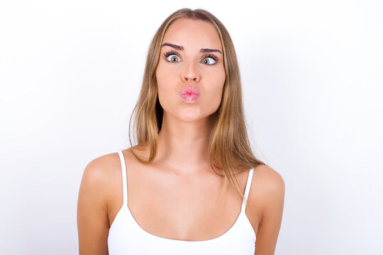 beautiful blonde girl wearing sexy t-shirt on white background crosses eyes, puts lips, makes grimace with awkward expression has fun alone, plays fool.