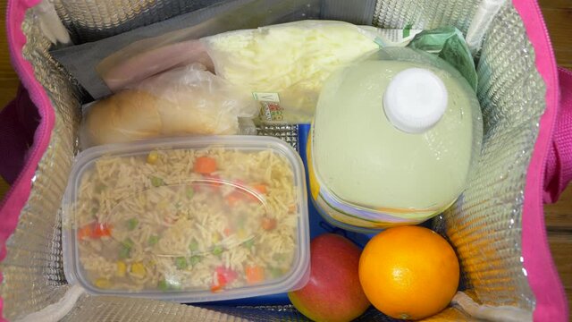 Closeup POV Overhead Shot Of A Man’s Hands Adding A Plastic Tub Of Vegetable Rice, Followed By Cutlery Wrapped In A Napkin, To An Insulated Cool Bag With Ice Blocks On The Bottom.