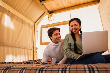 Multiethnic couple using gadgets on bed in glamping house