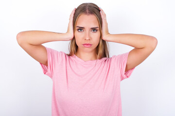 Fototapeta na wymiar Frustrated beautiful blonde girl wearing pink t-shirt on white background plugging ears with hands does not wanting to listen hard rock, noise or loud music.