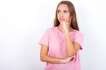 beautiful blonde girl wearing pink t-shirt on white background with hand under chin and looking sideways with doubtful and skeptical expression, suspect and doubt.
