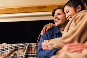 Low angle view of laughing interacial couple in blanket sitting on bench outdoors