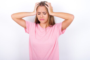 beautiful blonde girl wearing pink t-shirt on white background holding head with hands, suffering from severe headache, pressing fingers to temples