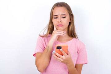 Thinking dreaming beautiful blonde girl wearing pink t-shirt on white background using mobile phone and holding hand on face. Taking decisions and social media concept.