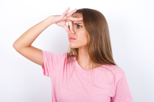 Displeased beautiful blonde girl wearing pink t-shirt on white background plugs nose as smells something stink and unpleasant, feels aversion, hates disgusting scent.