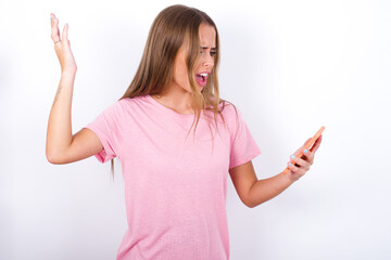 Photo of outraged annoyed beautiful blonde girl wearing pink t-shirt on white background holds cell phone, makes call, argues with colleague,  expresses negative emotions. People and anger.