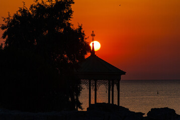 the silhouette of trees and a beautiful elegant gazebo with a cross on the roof against the background of a bright orange sunset sun and a calm sea