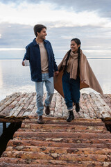 Smiling interracial couple with blanket and cup walking on pier near sea