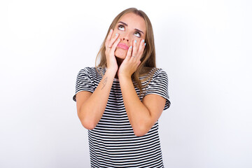 beautiful blonde girl wearing striped t-shirt on white background keeps hands on cheeks has bored displeased expression. Stressed hopeless model