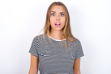 Fototapeta premium Shocked beautiful blonde girl wearing striped t-shirt on white background stares bugged eyes keeps mouth opened has surprised expression. Omg concept