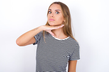 beautiful blonde girl wearing striped t-shirt on white background cutting throat with hand as...