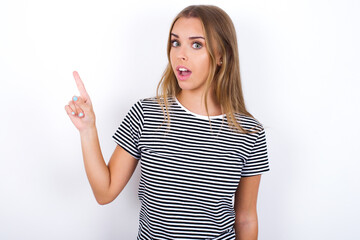 Emotive beautiful blonde girl wearing striped t-shirt on white wall keeps jaw dropped from shock demonstrates amazing promo points right on blank space demonstrates big shopping sale. Advertisement