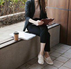Young millennial businesswoman sitting by the office building and drinking coffee