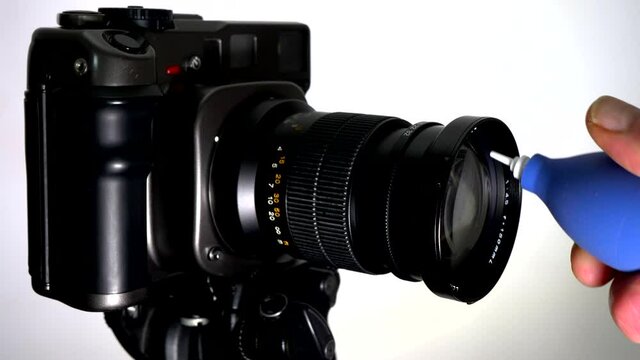 Closeup isolated shot of a man&rsquo;s hand removing the cap / cover of a medium format film camera lens on a tripod, then using a blue rubber blower to remove dust from the glass, before taking a picture.