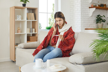 Sad woman sitting on sofa at home, looking at thermometer in hand and talking to doctor on phone....