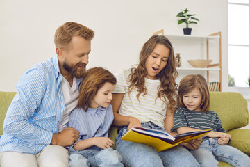 Happy family spending time together. Parents and kids sitting on sofa and reading a book. Beautiful young mother reading interesting stories to her children while sitting on couch at home