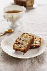 Slice of Pound Loaf Cake with raisins on white porcelain plate and cup of tea on greige linen tablecloth. Selective focus