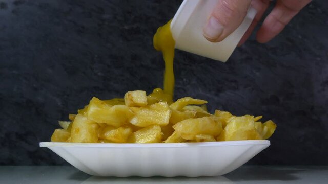 Closeup POV shot of a man&rsquo;s hand pouring thick hot curry sauce in a small tub, over a heaped portion of chips in a takaway / takeout polystyrene tray.