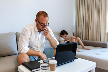Smiling dad working at home with his child on the sofa watching cartoons on tablet. Young single...