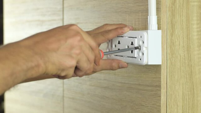 Electrician is using a screwdriver to install a power outlet in to a plastic box on a wooden wall.