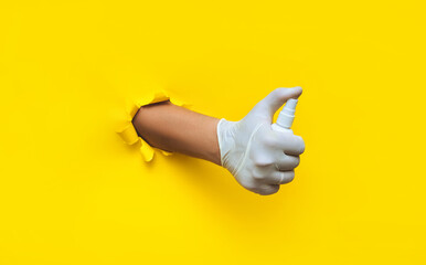 A right doctor's hand in a white medical glove holds a spray sanitizer. Torn hole in yellow paper, copy of the space. Concept of protection against coronavirus, covid-19 and other bacteria and viruses