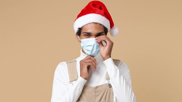 Young Santa curly african american man 20s in white shirt Christmas hat sterile face mask ppe to safe from coronavirus covid-19 on lockdown isolated on plain pastel beige background studio portrait