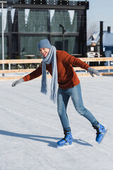 full length of happy young man in sweater, scarf and winter hat skating on ice rink