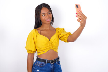 Portrait of a afro american woman with braids wearing sexy yellow t-shirt on white wall taking a selfie to send it to friends and followers or post it on his social media.