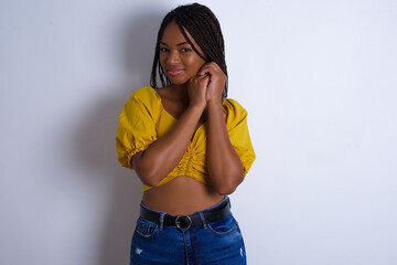 Charming serious afro american woman with braids wearing sexy yellow t-shirt on white wall keeps hands near face smiles tenderly at camera