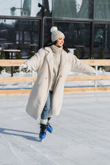 full length of happy young woman in winter hat and coat skating on ice rink
