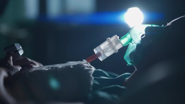 A female patient in medical mask is breathing oxygen support in a hospital next to heart rate machine. Unhealthy woman in coma. Getting mechanical ventilation. Concept of pandemic and therapy.
