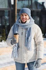 happy young man in winter hat and scarf holding paper cup outside