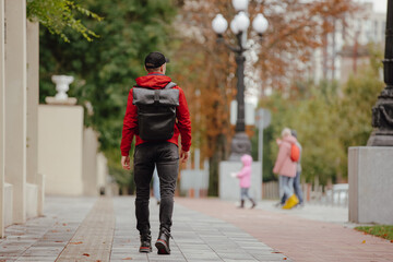 Back view man in red jacket with backpack walking along autumn alley