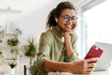 Young black woman using cellphone while working with laptop