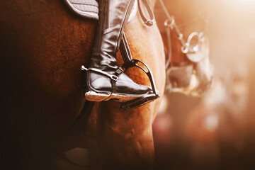 A rider in black boots with spurs sits on a bay racehorse in the saddle with stirrups, illuminated by sunlight. Equestrian sports. Horse riding.