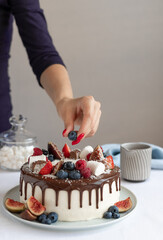 Woman decorating birthday cake with fresh berries and chocolate ganache. delicious homemade sweet baked dessert. gray wall background. vertical image