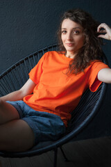 Fototapeta na wymiar Female portrait of a beautiful curly girl in bright orange t-shirt sitting at home in her apartment on dark background, happy people concept