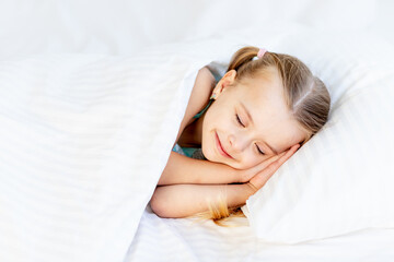 Obraz na płótnie Canvas a girl child sleeps on a bed at home on a white cotton bed and smiles sweetly in her sleep with her hands folded under her cheeks