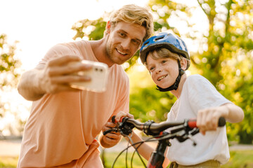 Obraz premium Ginger man taking selfie while teaching his son how to ride bicycle