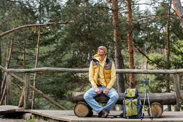 traveler looking away while resting on log bench near backpack and trekking poles