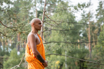 side view of tattooed man in traditional buddhist robe meditating in forest