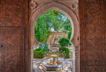 Puerta árabe abierta hacia un hermoso jardín con fuentes, estanques y vegetación.