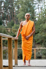 full length view of buddhist monk standing near wooden fence in forest - obrazy, fototapety, plakaty