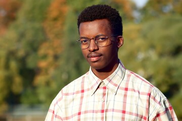 Portrait of serious handsome black African Afro American attractive guy student in glasses and shirt looking at camera, young ethnic man standing, walking in golden autumn park.
