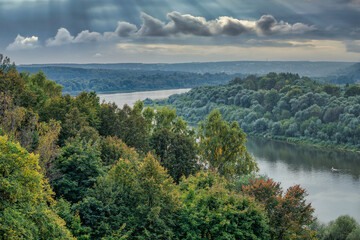 Obraz premium A high altitude shot of a river flowing along the banks overgrown with trees and lush grass against the sunset sky. Rural landscape in autumn, city structures in the background.