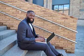 A dark-skinned man in a suit sitting on the steps with a laptop