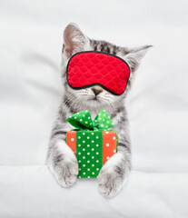 Cozy scottish kitten holds gift box and sleeps in sleeping mask under blanket on a bed at home. Top...